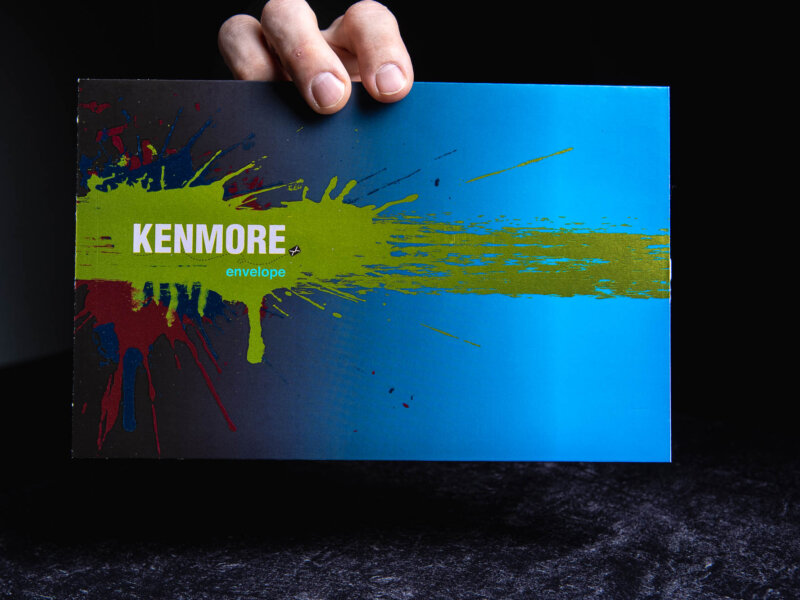 Kenmore Envelope logo with a splattered yellow, blue and red paint background