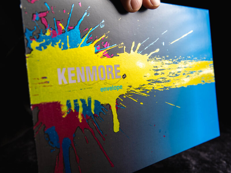 Kenmore Envelope logo with a splattered yellow, blue and red paint background