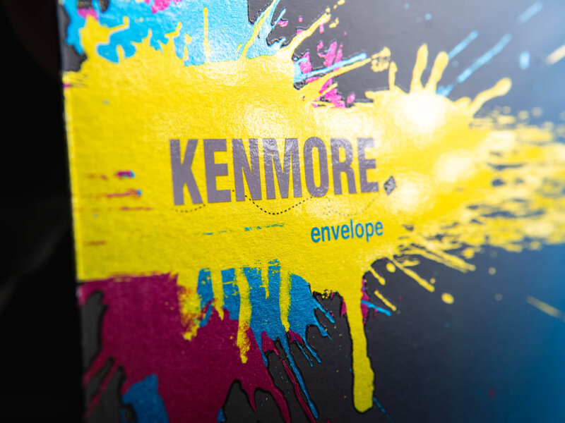 Kenmore Envelope logo with a splattered yellow, blue and red paint background